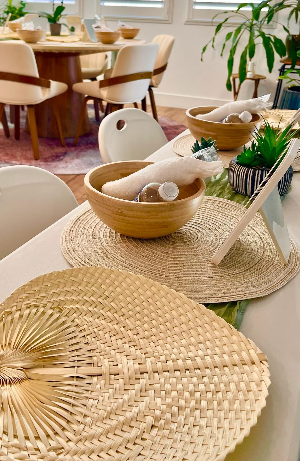 One guest's place setting: a bamboo bowl holding a rolled towel and bottled water, on a woven mat beside a small potted succulent and a mirror