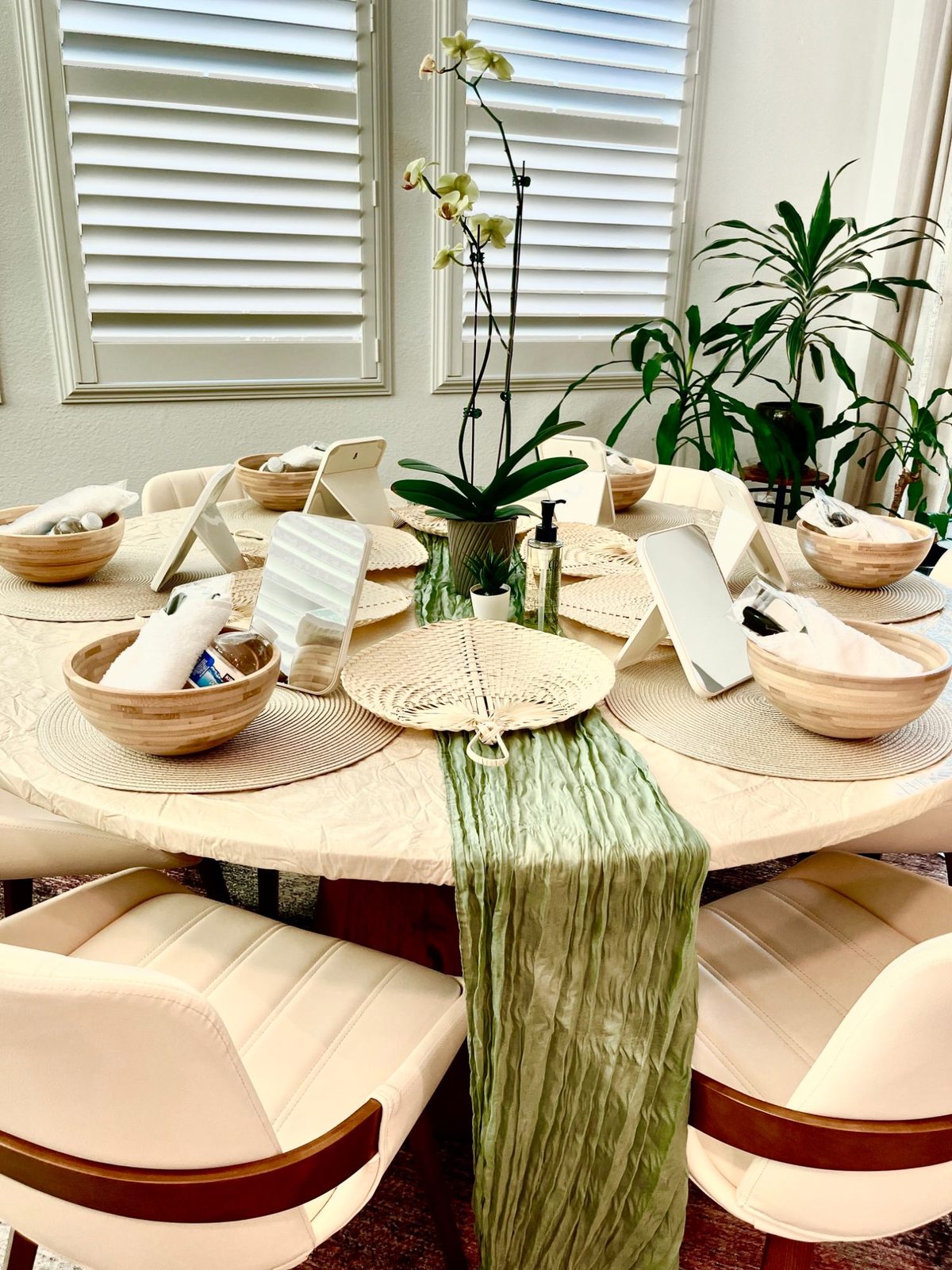 A round table set for a glow party: woven placemats, bamboo bowls holding rolled white towels, a standing mirror at every seat, an orchid at the centre and a sage green runner down the middle